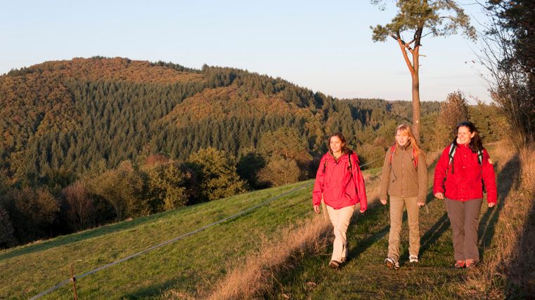 Three women walk along a path through an autumnal landscape with wooded hills in the background.
