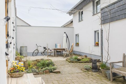 Ein gemütlicher Innenhof mit einer Holzbank und einem Tisch. Im Hintergrund steht ein Fahrrad und es gibt einige Pflanzen und Blumen.