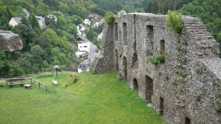 Eine beeindruckende Burgruine auf einer Wiese, umgeben von grünen Hügeln. Im Hintergrund sind Häuser in einer malerischen Landschaft zu sehen.
