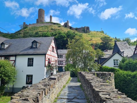 Eine malerische Landschaft mit einem historischen Burgturm im Hintergrund. Im Vordergrund führen Steine und Häuser mit Fachwerkarchitektur zu der Burg.