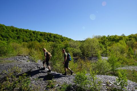 Two people walk through a green landscape with slate soil and trees under a clear blue sky.