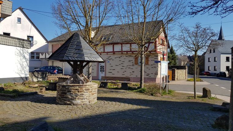 Ein malerischer Dorfplatz mit einem Brunnen in der Mitte. Alte Fachwerkhäuser und Bäume umgeben die Szenerie.