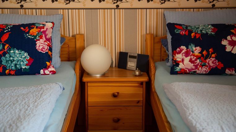 Ein gemütliches Schlafzimmer mit zwei Einzelbetten und bunten Kissen. Auf dem Nachttisch steht eine runde Lampe und eine kleine Uhr.