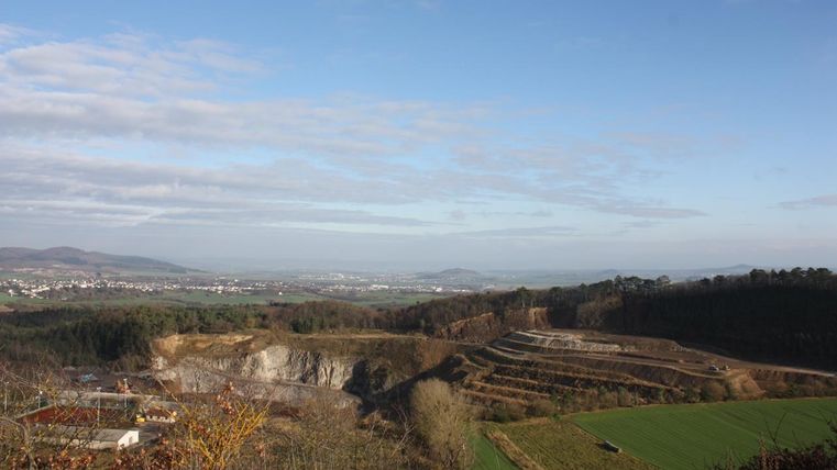 A wide landscape with gentle hills and green fields. In the background, trees and a clear sky are visible.