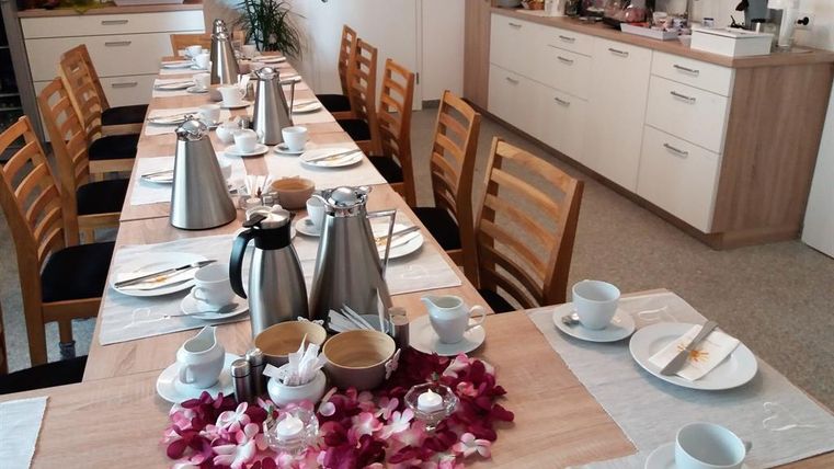 A set table with several coffee pots and cups, surrounded by chairs. In the background, a kitchenette with various treats is visible.