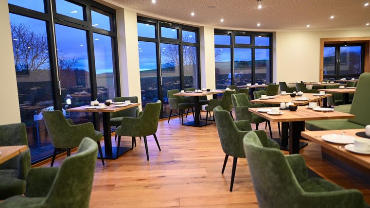 Ein modernes Restaurant mit großen Fenstern und gemütlichen, grünen Stühlen. Die Tische sind stilvoll gedeckt und der Holzfußboden verleiht eine warme Atmosphäre.