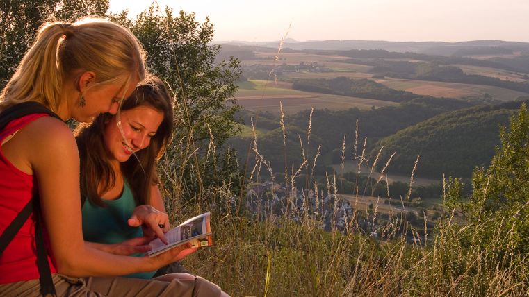 Zwei Frauen sitzen auf einem Hügel und schauen in ein Buch, im Hintergrund eine weite Landschaft mit Feldern und Hügeln.