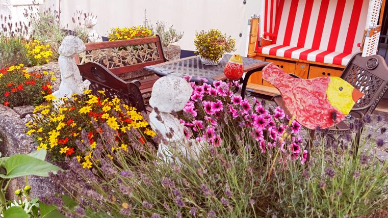 Ein schöner Garten mit bunten Blumen und Vogeldekorationen. Im Hintergrund steht ein Strandkorb mit rot-weißem Streifenmuster.