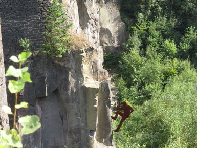 Ein Kletterer steht an einer steilen Felswand, umgeben von üppigem Grün. Die Szene vermittelt ein Gefühl von Abenteuer und Naturverbundenheit.