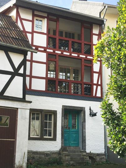Ein charmantes Fachwerkhaus mit roten Balken und einer türkisfarbenen Haustür. Umgeben von Pflanzen und einem klaren Himmel.