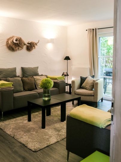 A modern living room with cozy furnishings and a table in the center. The walls are bright, and there is a view of the outdoor area through the windows.