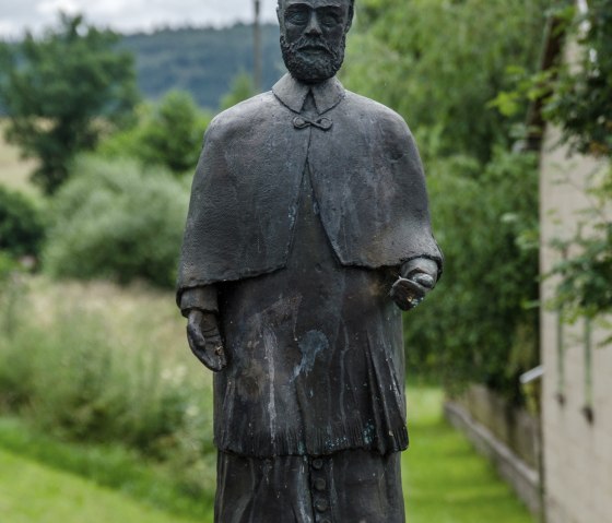 Statue en bronze d'un homme en tenue traditionnelle sur un socle, entour&eacute; d'un paysage verdoyant et d'un ciel nuageux., &copy; Volker Windheuser