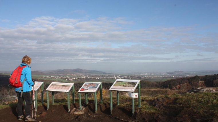 A person stands in front of information signs and looks over a landscape. The sky is slightly cloudy and it is a beautiful vantage point.