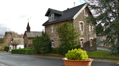 Ein charmantes Steinhaus mit einem schwarzen Dach und einem kleinen Garten. Im Hintergrund ist eine Kirche mit einem schmalen Turm zu sehen.