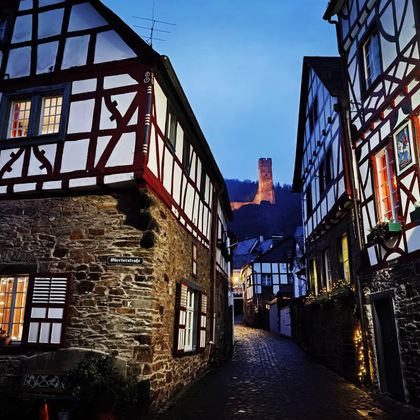 Eine malerische Gasse mit Fachwerkhäusern und warmem Licht. Im Hintergrund ist eine Burg auf einem Hügel sichtbar.