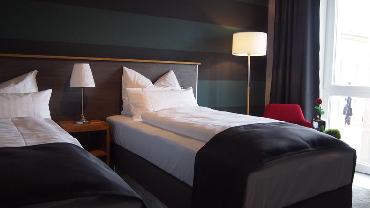 A modern hotel room with two single beds and a large window. The room is decorated with a floor lamp and a small table.