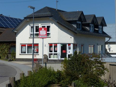 Ein modernes Gebäude mit mehreren Fenstern und einem roten Logo. Vor dem Haus befinden sich grüne Pflanzen und ein Gehweg.