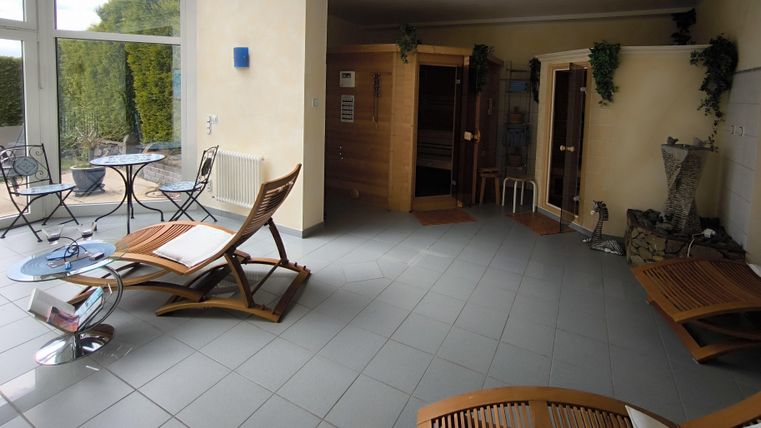 Ein heller Entspannungsraum mit Liegestühlen und einem Blick auf eine Sauna. Die Wände sind mit Pflanzen dekoriert, und der Raum hat große Fenster.