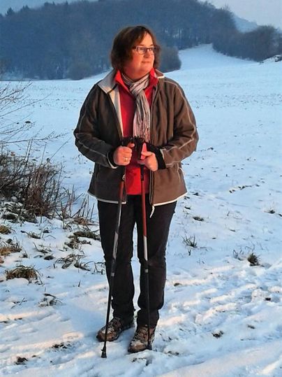 A person is standing in a snowy field holding two hiking poles. In the background, trees and gentle hills are visible.
