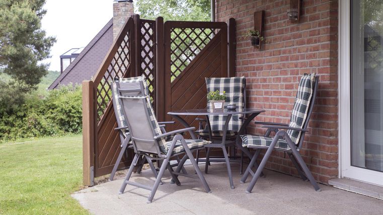 A cozy outdoor area with a table and four chairs on a terrace. In the background, there are plants and a wooden wall.