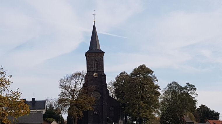 Eine alte Kirche mit einem hohen, spitzen Turm steht zwischen Bäumen. Der Himmel ist leicht bewölkt und strahlt eine ruhige Atmosphäre aus.