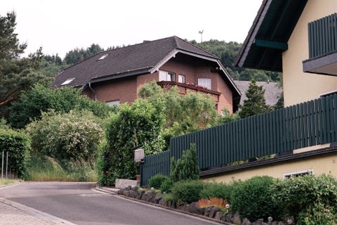Eine ruhige Wohnstraße mit zwei Häusern und viel Grün. Die Umgebung ist von Bäumen und Sträuchern umgeben.