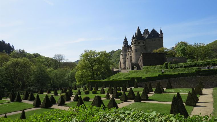 Ein beeindruckendes Schloss umgeben von gepflegten Gärten mit geometrischen Hecken. Die Landschaft ist grün und sonnig, ideal für einen Spaziergang.