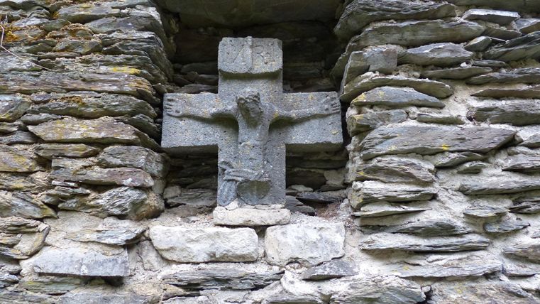 A cross made of stone, embedded in a wall of slate stones. The texture of the stones is rustic and natural.
