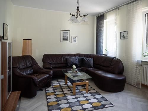 Ein modernes Wohnzimmer mit einer großen, braunen Ledercouch und einem Glastisch. Der Raum ist hell und freundlich, mit einem bunten Teppich und Kunst an den Wänden.