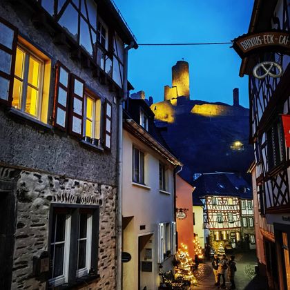 Eine malerische Gasse mit Fachwerkhäusern und festlicher Beleuchtung. Im Hintergrund ragt eine beleuchtete Burg in den Abendhimmel.