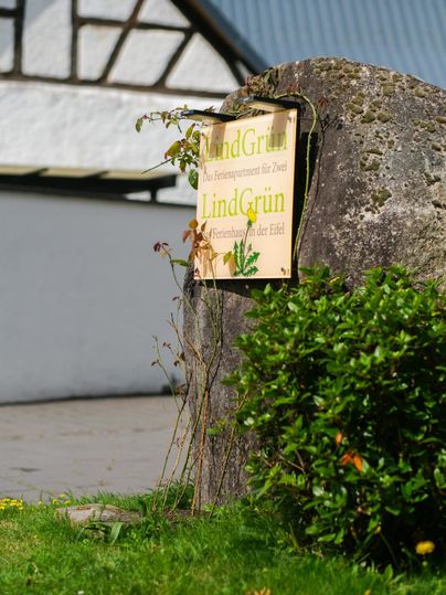 A sign that reads "LindGrün" stands by a large stone. In the foreground, a green bush can be seen.