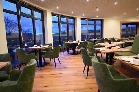 Ein modernes Restaurant mit großen Fenstern und gemütlichen, grünen Stühlen. Die Tische sind stilvoll gedeckt und der Holzfußboden verleiht eine warme Atmosphäre.