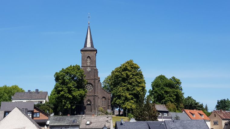 Eine schöne Kirche mit einem hohen Turm steht über den Dächern der Stadt. Umgeben von grünen Bäumen und klarem blauen Himmel.