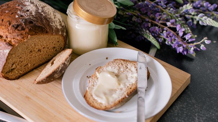 Fresh bread with butter on a plate, accompanied by a glass of cream-colored butter. In the background, there are purple flowers visible.