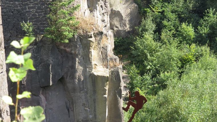 Eine Person klettert an einer Felswand in einer grünen Umgebung. Im Hintergrund sind Bäume und eine steil abfallende Klippe sichtbar.