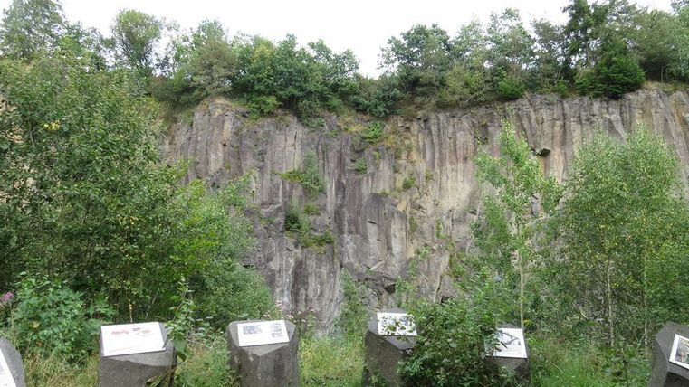 Ein steiler Felsen mit grüner Vegetation im Vordergrund. Informationsschilder sind entlang des Weges aufgestellt.