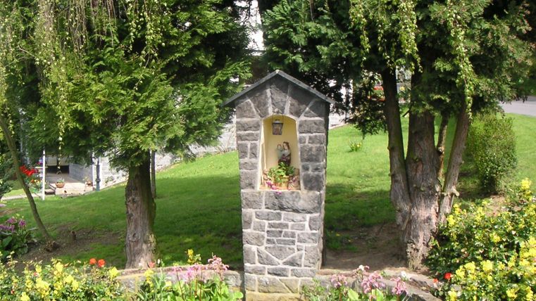 A small stone column surrounded by colorful flowers and trees. In the column stands a statue.