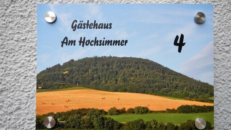A sign with the inscription "Gästehaus Am Hochsimmer" and the number 4. In the background, a green hilly landscape with fields can be seen.