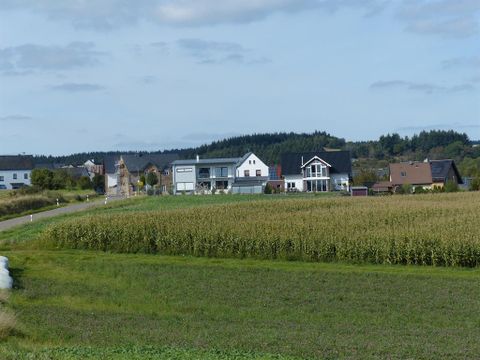 Eine ländliche Szene mit Häusern, die von Feldern umgeben sind. Im Hintergrund sind sanfte Hügel und ein klarer Himmel zu sehen.