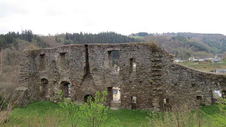 An old castle ruin surrounded by green grass and trees. In the background, gentle hills and a small village can be seen.