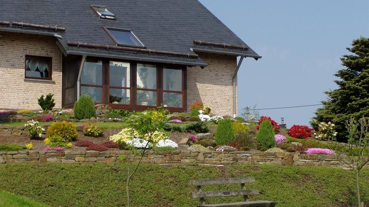 A lovely house with a well-maintained garden full of flowers. In the foreground, there is a bench on the lawn.