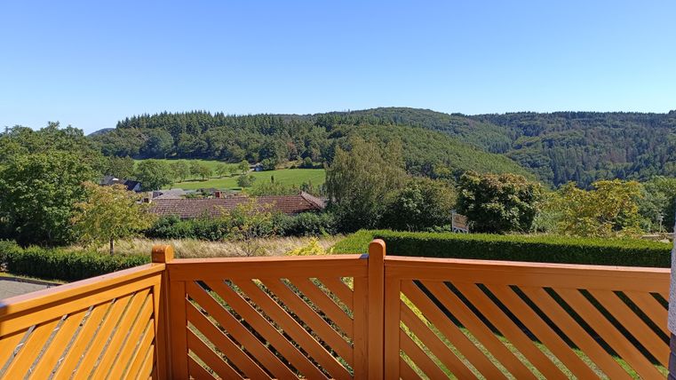 Eine schöne Aussicht von einer Terrasse mit einem hölzernen Geländer. Im Hintergrund sind grüne Hügel und eine klare, blaue Himmel zu sehen.