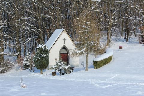 Eine kleine Kapelle im Schnee, umgeben von Bäumen. Die Landschaft ist friedlich und winterlich.