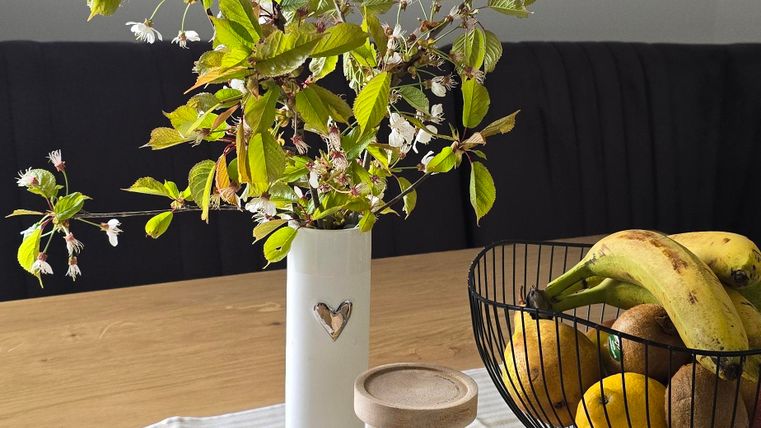 Ein Tisch mit einer Vase, die frische Zweige mit Blättern zeigt. Daneben stehen eine Obstschale mit Bananen und ein Glas mit Dekoration.