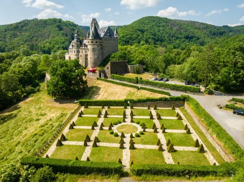 An impressive castle surrounded by green hills. In the foreground, there is a geometric garden with intricately designed hedges.