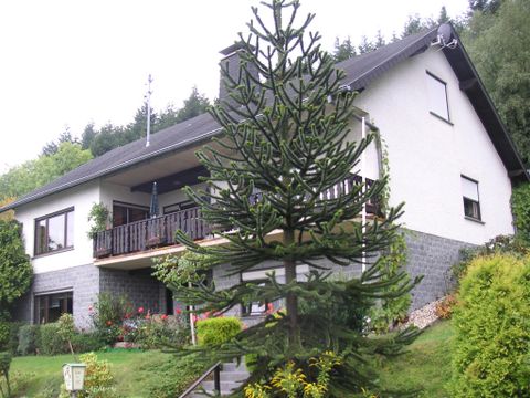 Ein zweistöckiges Haus mit einem Balkon und einem schönen Garten. Im Vordergrund steht ein hoher Baum.