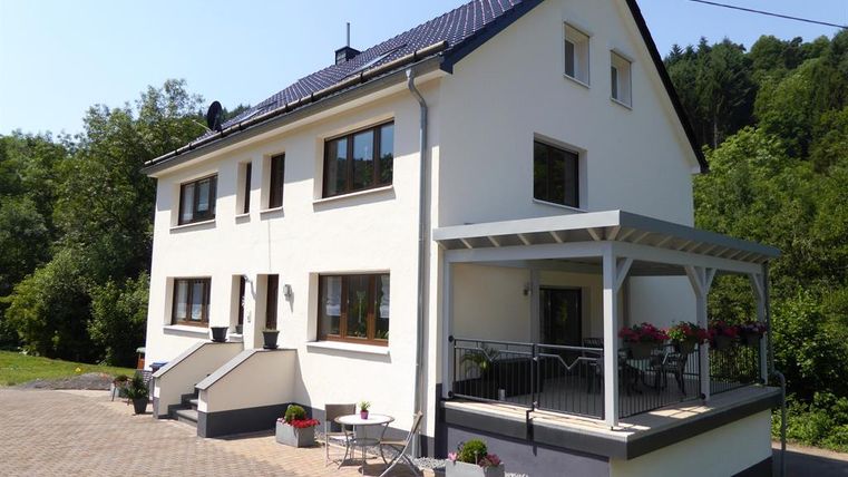 Ein modernes Haus mit einem großen Balkon und einer Terrasse. Umgeben von Grün und einem gepflasterten Gartenbereich.