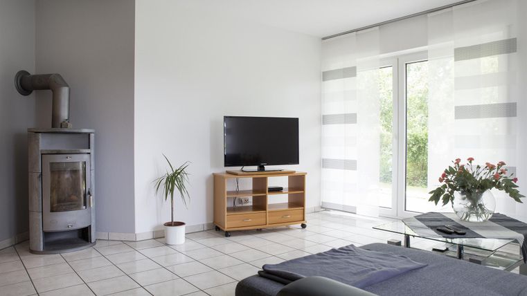 A modern living room with a wood stove, a television, and a wooden shelf. Large windows let in plenty of light and there are plants on the table.