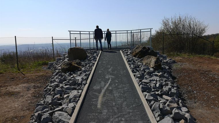 Ein Weg aus Steinen führt zu einer Aussichtsplattform. Zwei Personen stehen am Ende des Weges und blicken in die Ferne.