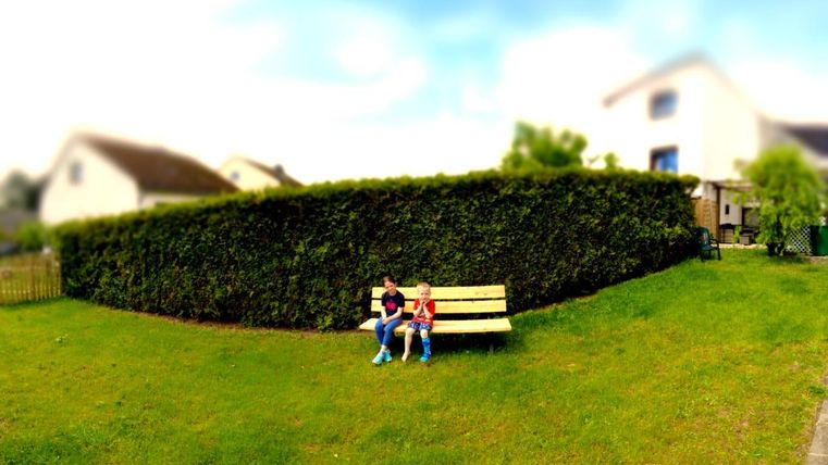 Zwei Kinder sitzen auf einer Bank im Garten. Im Hintergrund sieht man eine Hecke und einige Häuser.